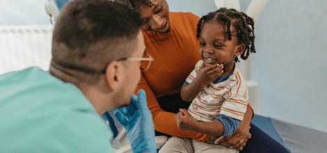 young male dentist interacting with small toddler who is sitting on his mothers lap smiling and facing the dentist 